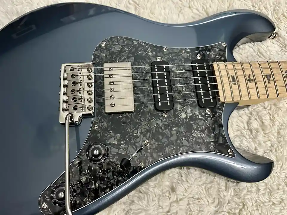 A close-up photo of a blue electric guitar with a black, cracked pickguard, silver hardware, and a light wood fretboard…