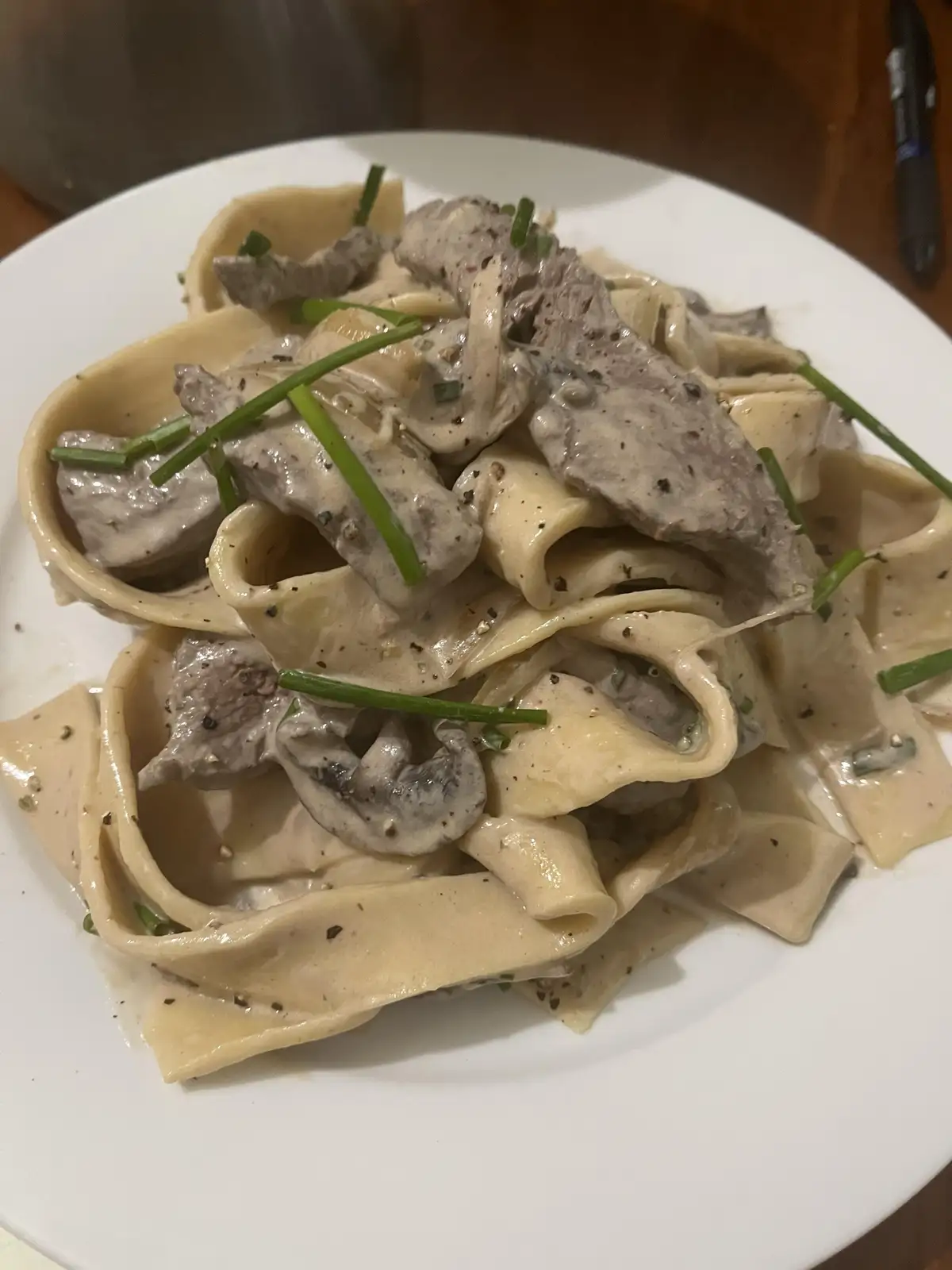 A close-up photo of a white plate holding wide pasta noodles covered in a creamy sauce, topped with sliced meat, mushrooms…