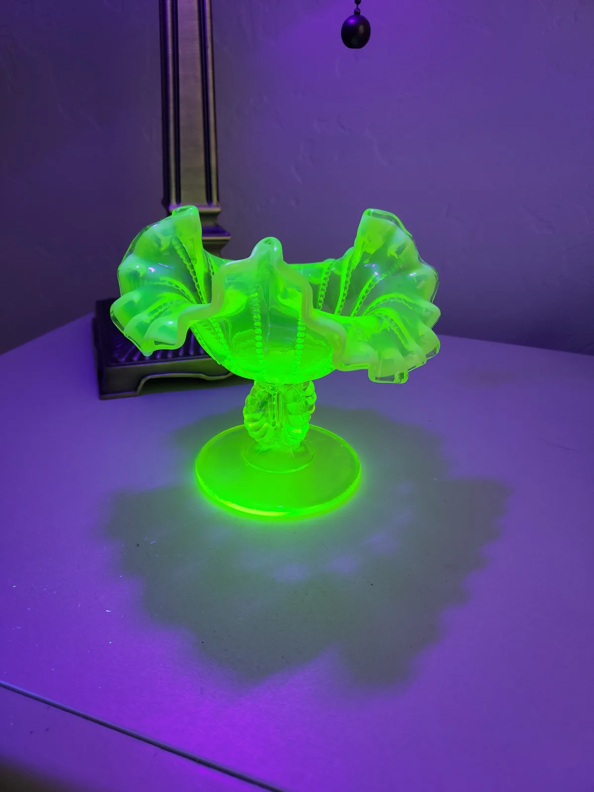 A vintage, ruffled glass dish with a pedestal base glows bright neon green under ultraviolet light, casting a vivid shadow…