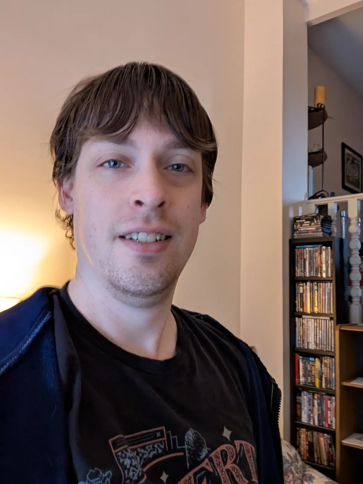 A man with brown hair and blue eyes smiles slightly while wearing a black t-shirt and a dark blue jacket, standing indoors…