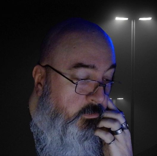 A man with a gray beard and glasses, eyes closed, hand near his mouth, appears contemplative or annoyed under blue…