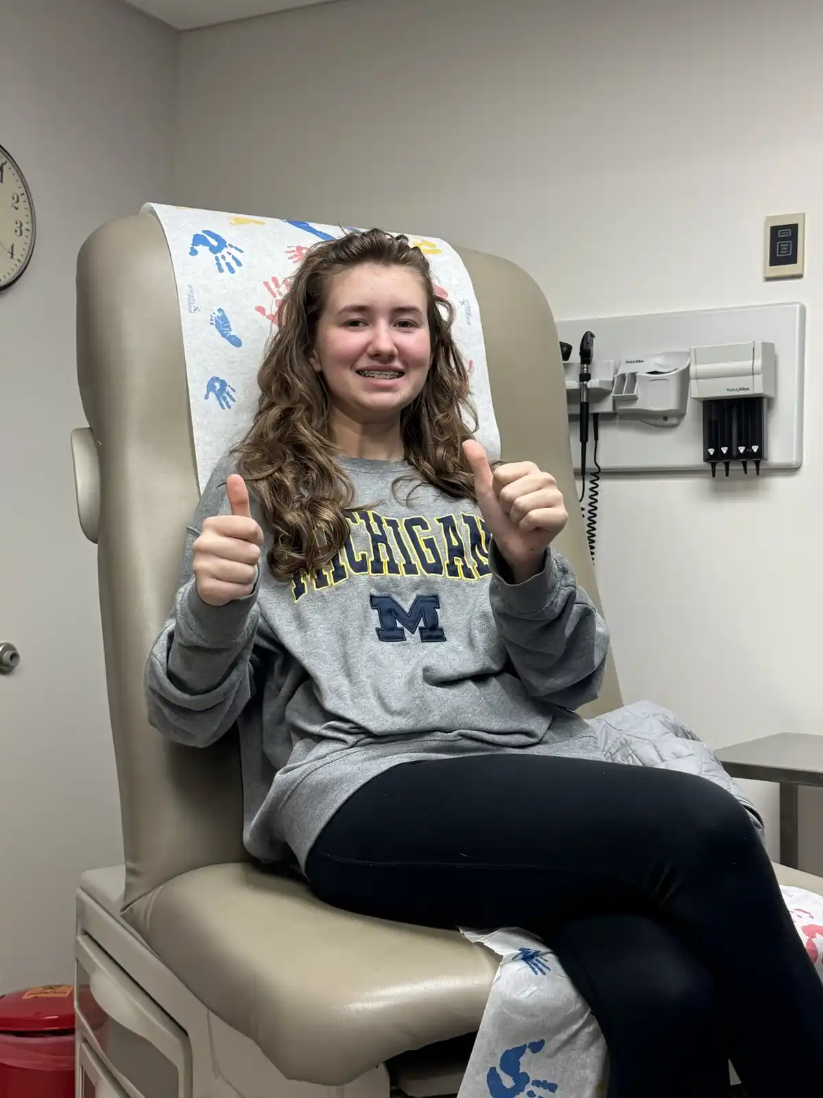 A young woman with braces sits in a medical exam chair, smiling broadly and giving two thumbs up. She wears a gray Michigan…