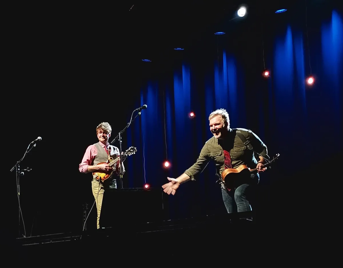 Two male musicians are on a dimly lit stage with blue and red spotlights. The man on the right, holding an acoustic guitar…