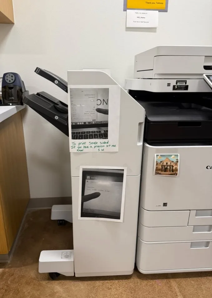 A white office printer has two printed images taped to its front panel. One image shows a laptop screen with a finger…