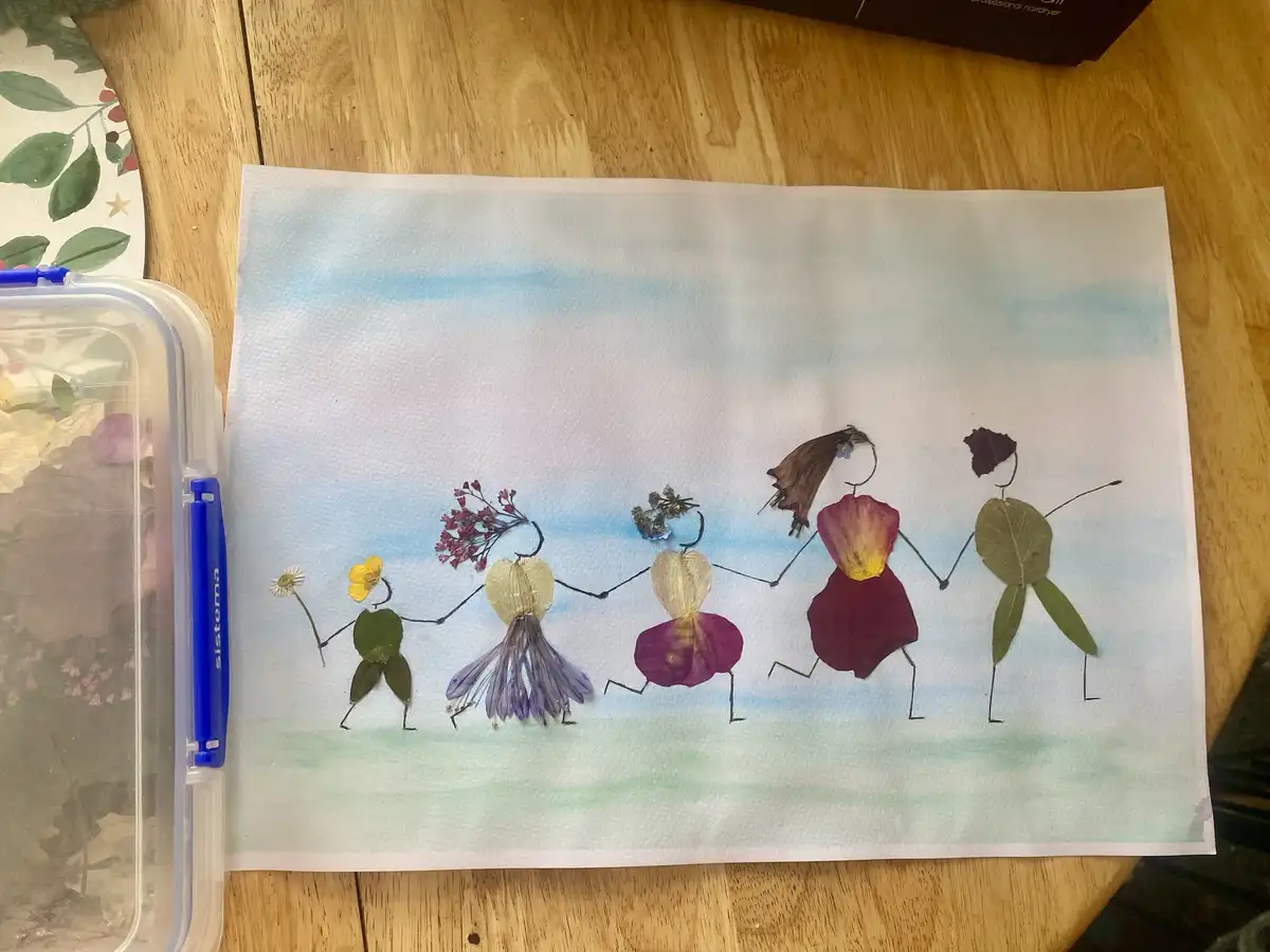 A handmade artwork on paper depicts five stick-figure people holding hands, each constructed from different pressed flowers…
