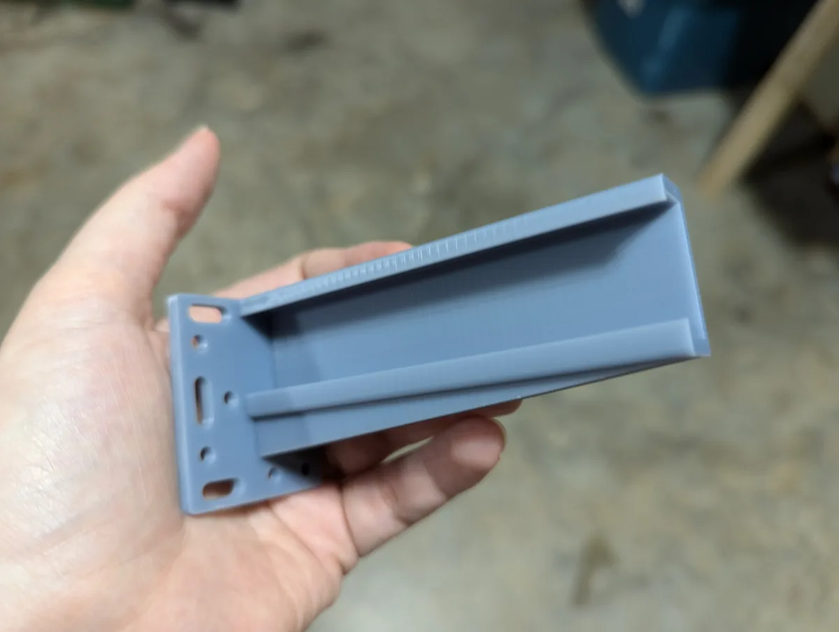 A hand holds a light blue 3d-printed plastic bracket with visible layer lines and mounting holes, against a blurred concrete…