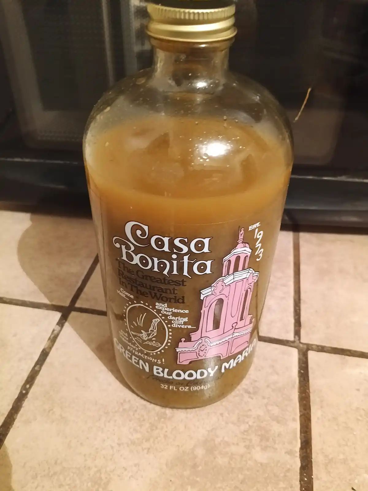 A bottle of Casa Bonita Green Bloody Maria sits on a tiled floor, its label featuring a pink illustrated building and text…