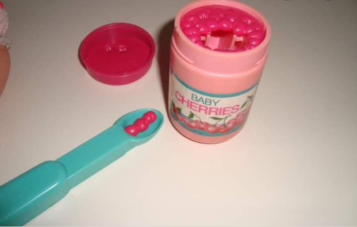 A pink plastic container labeled 'BABY CHERRIES' with a matching pink lid beside it, both sitting on a white surface. A teal…