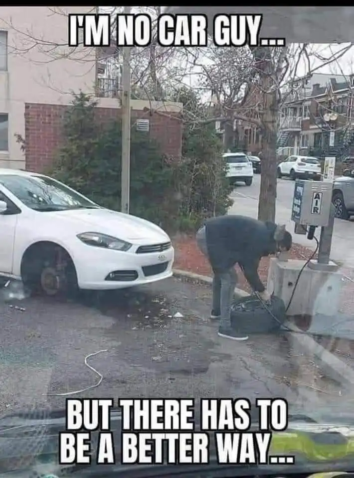 A person in a dark hoodie and jeans is bent over, manually inflating a tire using an air compressor hose connected to a…