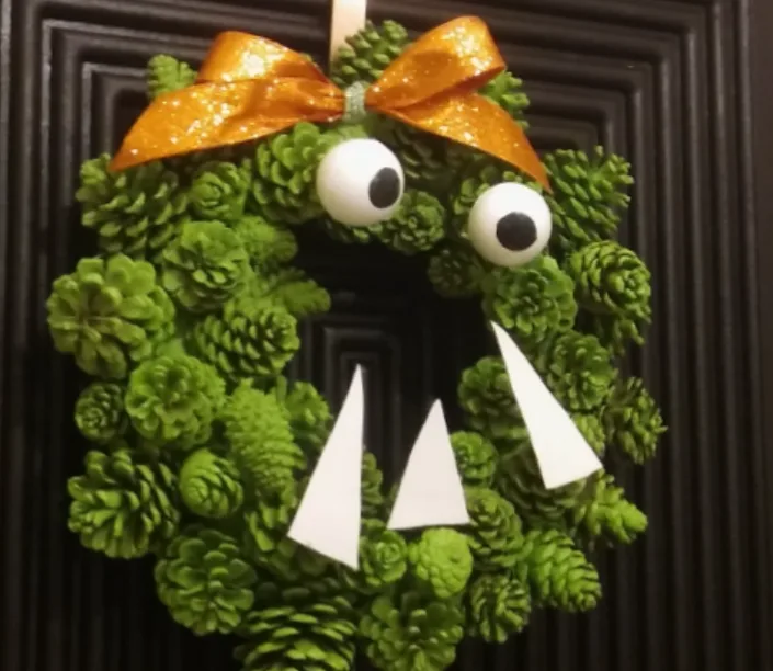 A green pinecone wreath is decorated to look like a monster, featuring two large googly eyes, a glittery orange bow on top…