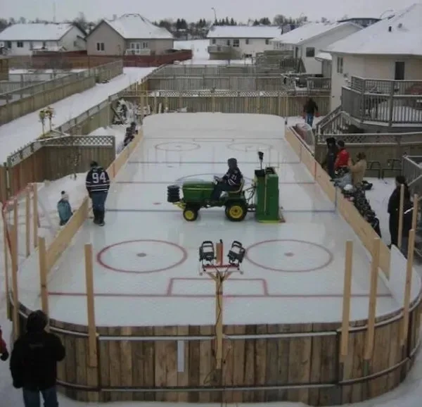 An aerial view of a backyard ice rink where a person in a hockey jersey drives a green John Deere tractor across the ice…