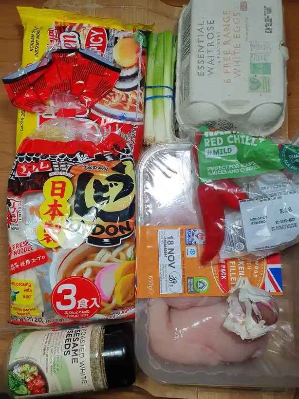 A flat-lay photo of grocery items including chicken fillets, red chillies, green onions, eggs, ramen noodles, and sesame…