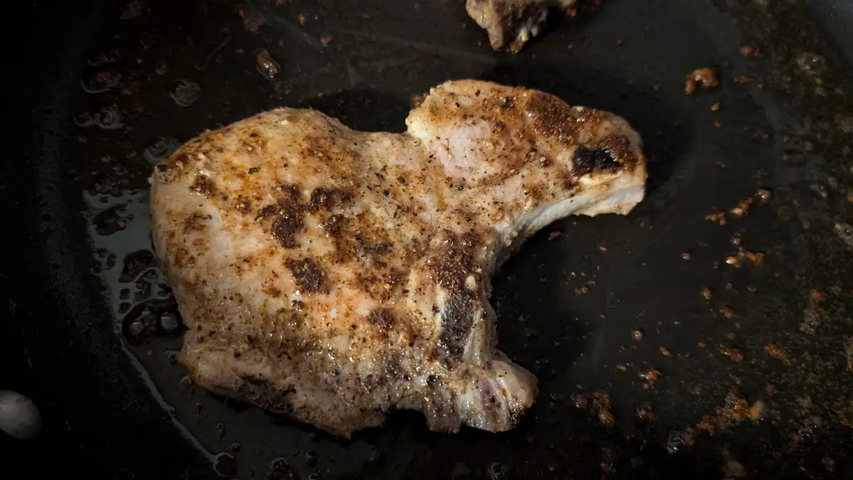 A close-up photo shows a seasoned pork chop sizzling in a dark, greasy frying pan. The meat has browned, charred spots from…