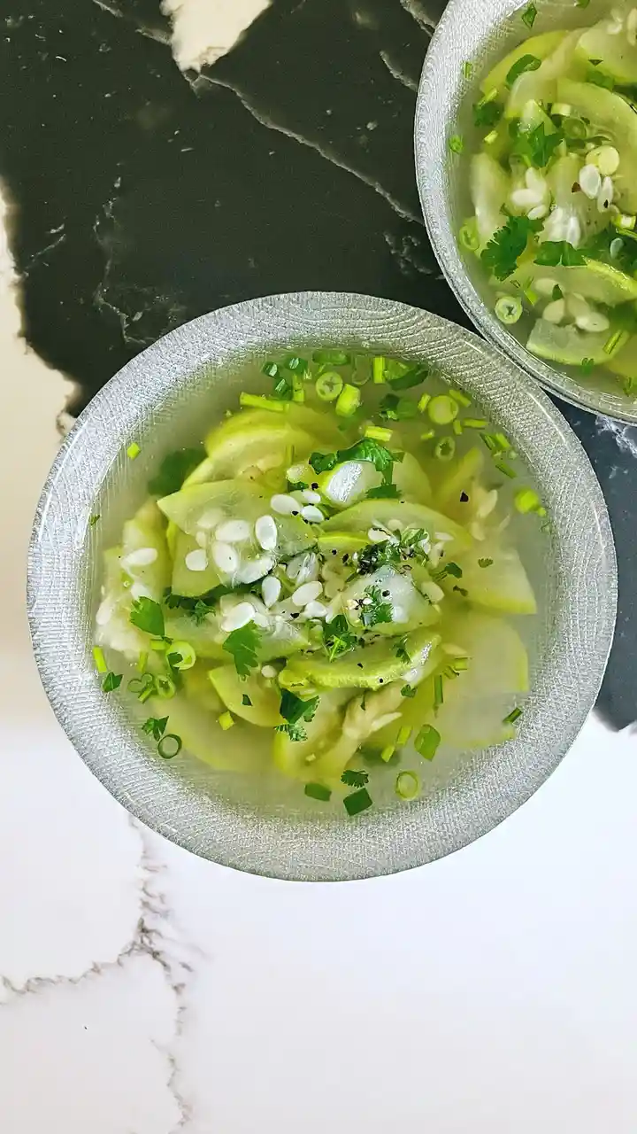 Two bowls of clear broth with sliced green vegetables, garnished with chopped green onions, cilantro, and white sesame…