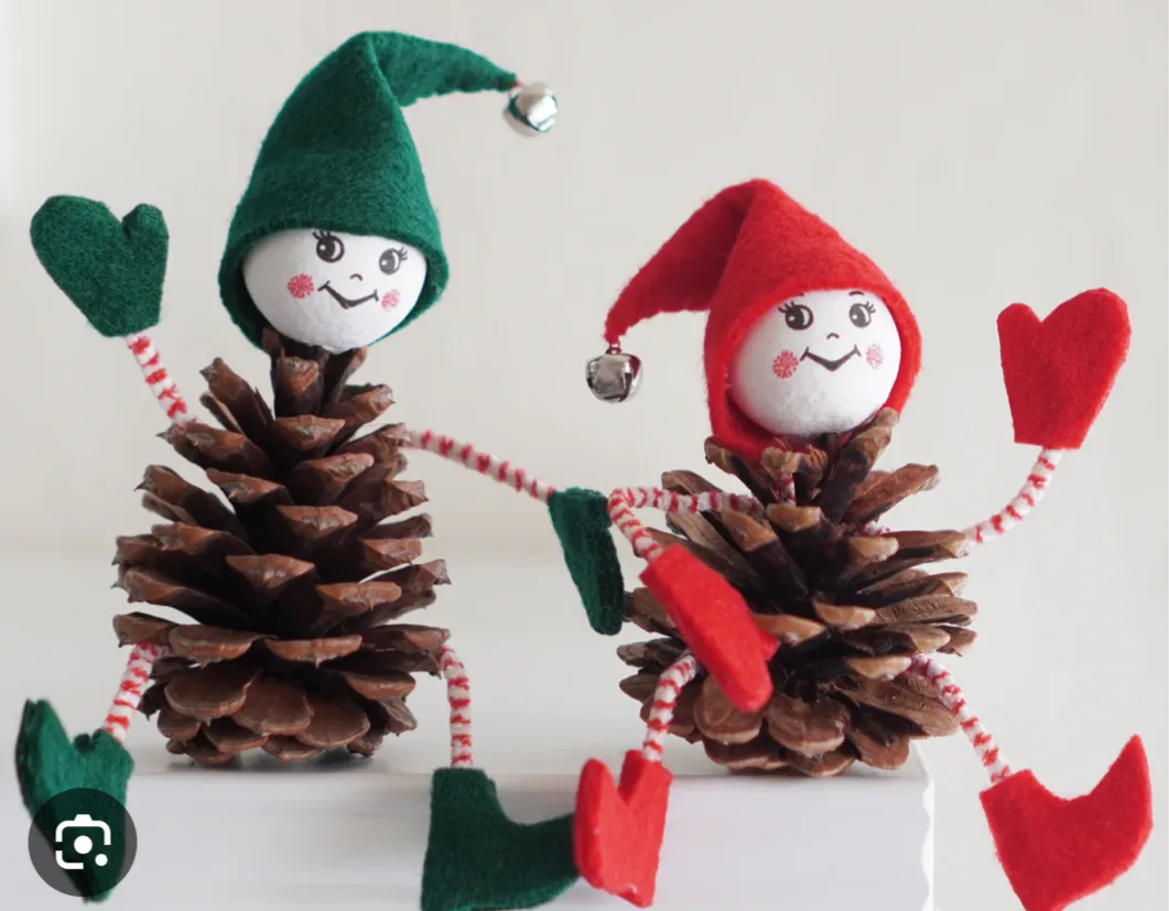 Two handmade pinecone elves, one in green and one in red, are posed holding hands with heart-shaped mittens. Each has a felt…