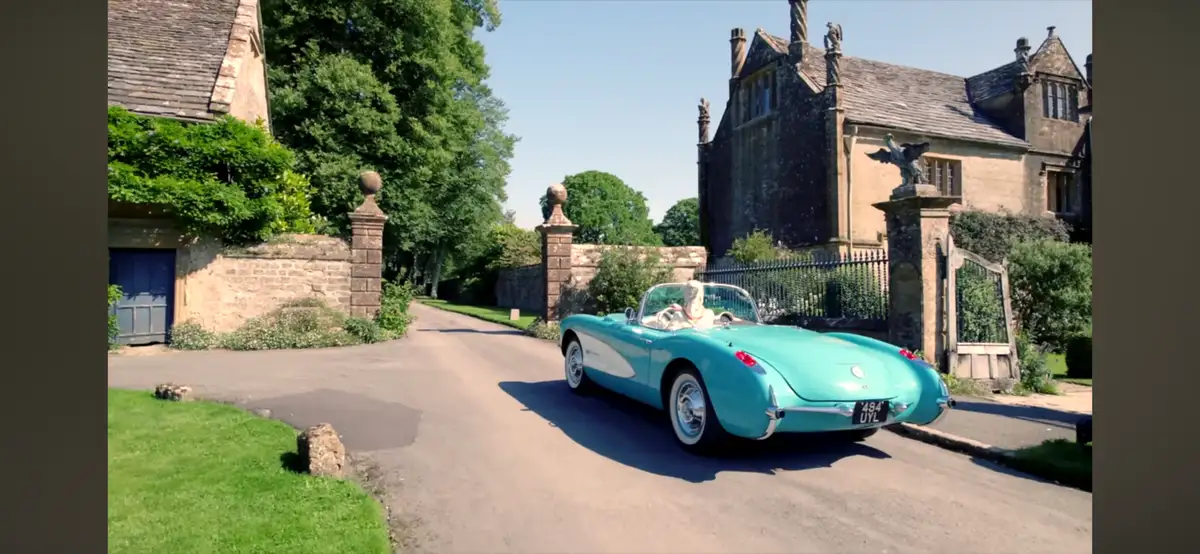A turquoise classic convertible with a white interior drives down a paved driveway toward a large stone manor house with…
