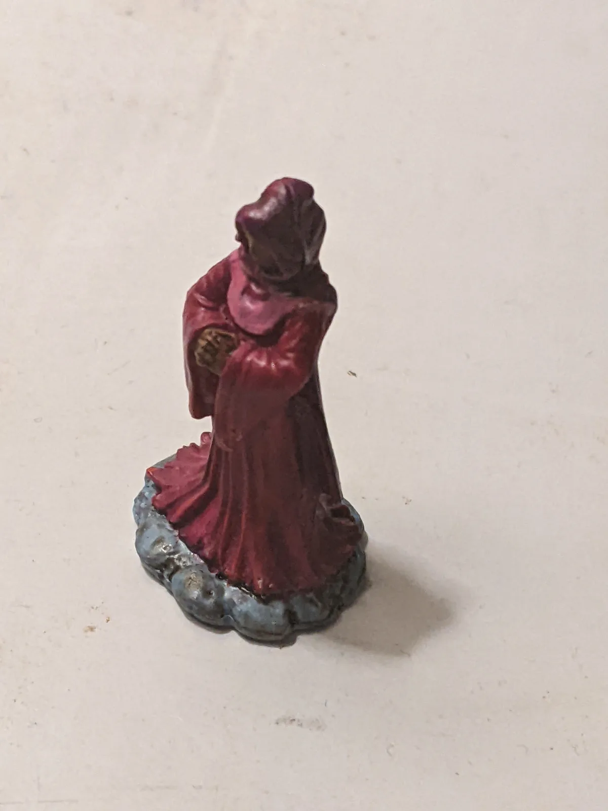 A small, painted figurine of a robed person in a deep red gown stands on a grey, scalloped base against a plain off-white…