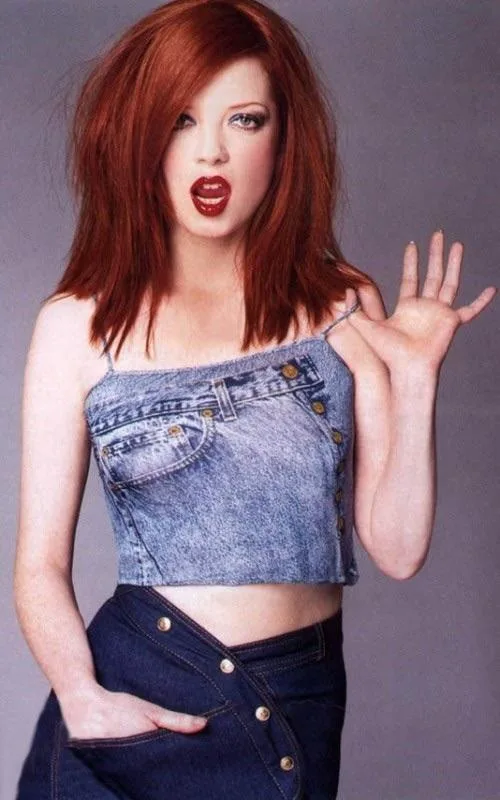 a woman with red hair and bold red lipstick wears a denim crop top and denim skirt, her mouth open in a surprised or…