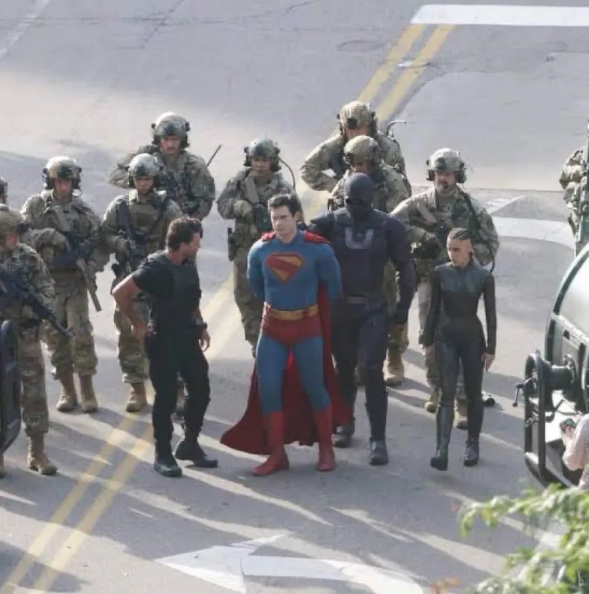 A behind-the-scenes photo shows actor Henry Cavill in a Superman costume being escorted by armed soldiers on a paved road…