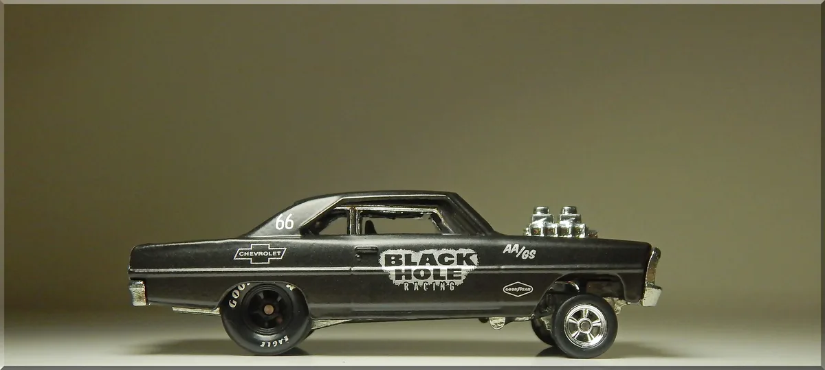 A black die-cast model of a classic Chevrolet race car, shown in profile against a plain background. The car features 'BLACK…
