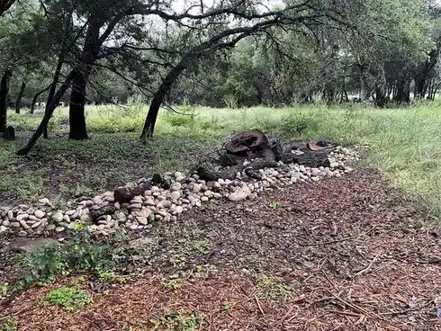 An outdoor scene shows a pile of cut logs and smooth stones arranged in a rough circle in a grassy, wooded area. The ground…