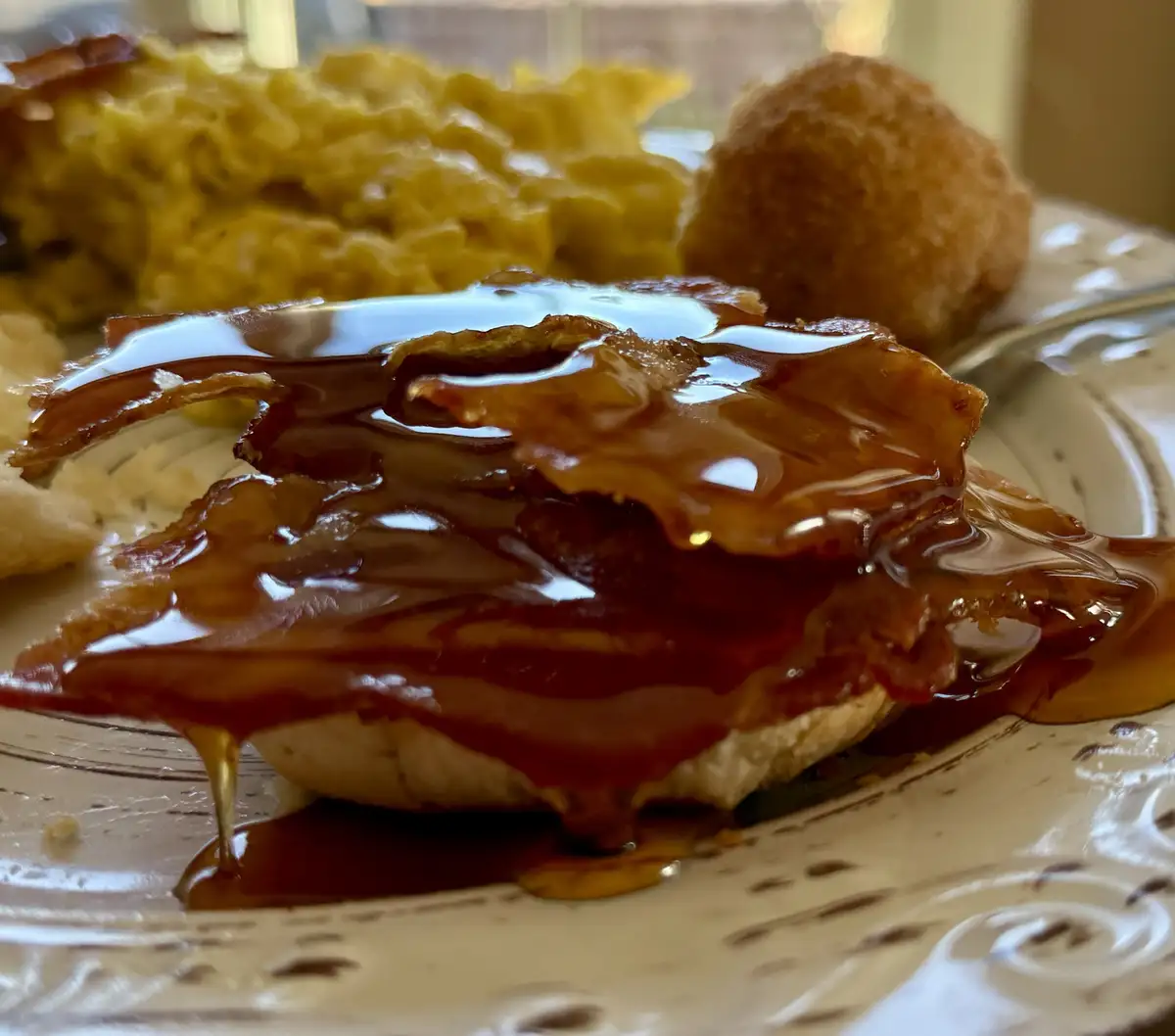 A close-up photo of a plate of food featuring a piece of bacon covered in a thick, glossy brown syrup, with macaroni and…