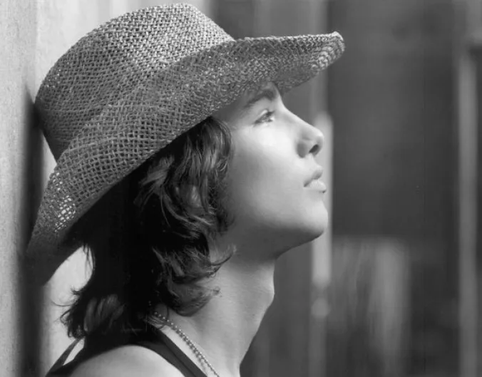A black-and-white photo shows a young person with wavy hair wearing a woven straw hat, looking upward with a serene…