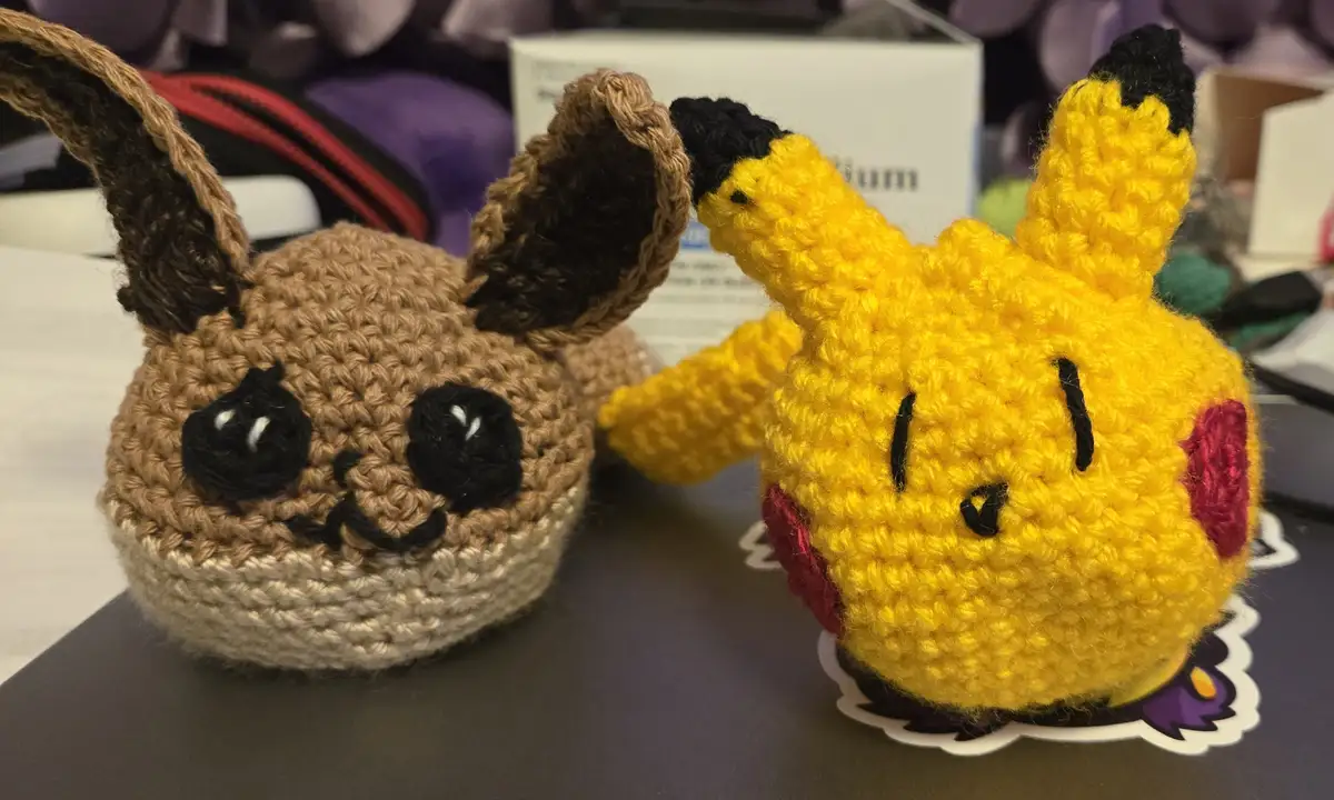 Two crocheted Pokémon plushes, a brown Eevee and a yellow Pikachu, sit side by side on a desk. Pikachu has its eyes closed…