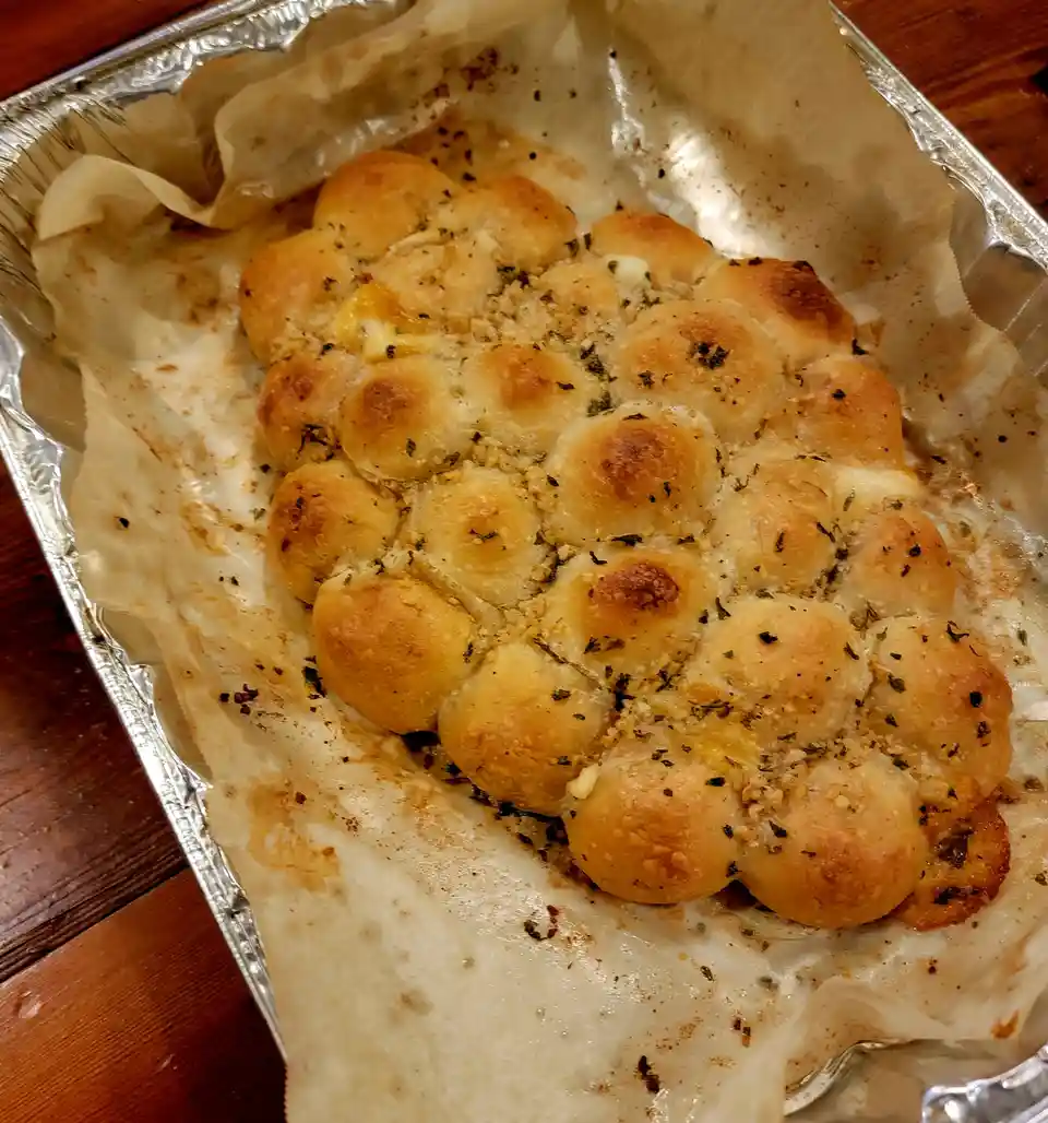 A close-up photo shows a baked garlic bread in a foil pan, featuring golden-brown, rounded, bubble-like dough pieces…