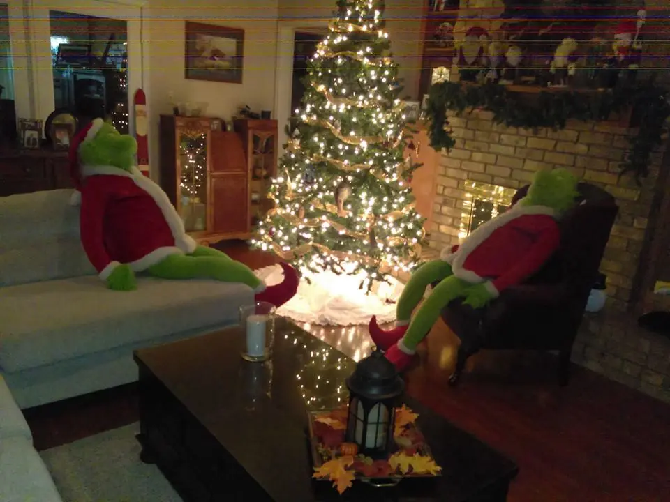 Two green Grinch plush toys, dressed in red Santa Claus outfits with white trim, are seated in a cozy living room. One sits…
