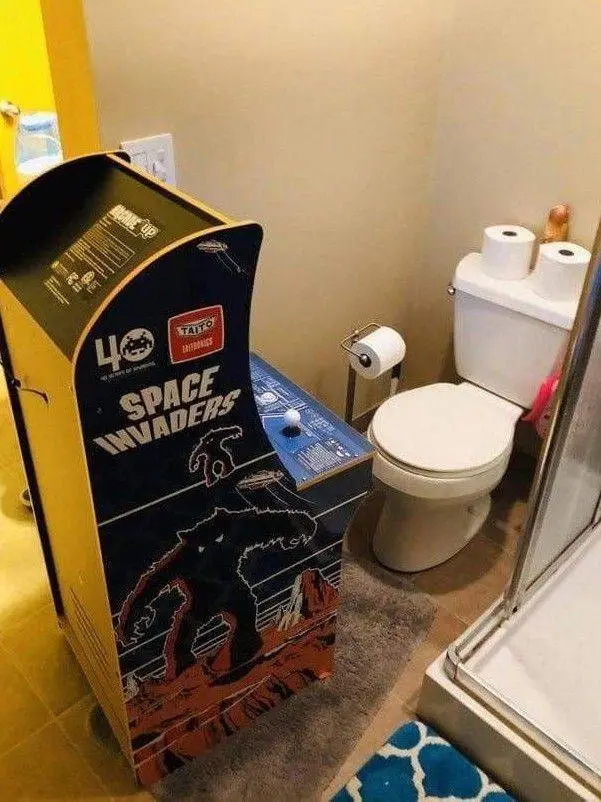 A bathroom with a toilet, shower, and a large arcade cabinet labeled 'Space Invaders' standing next to the toilet. The…