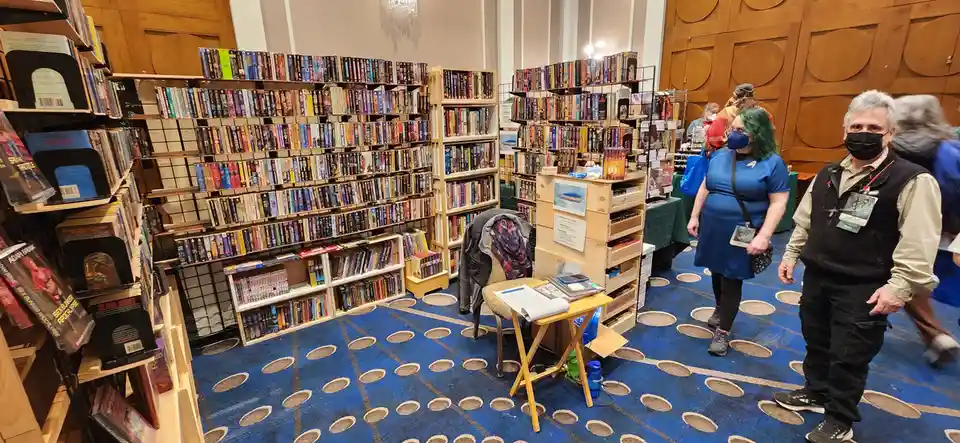 A photo of a crowded bookstore or convention floor filled with shelves of books and people browsing. Two people in the…
