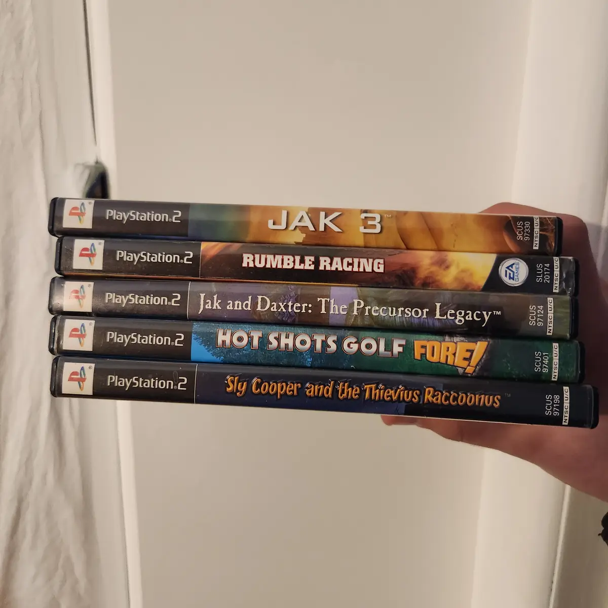 A hand holds five PlayStation 2 game cases vertically, showing titles including Jak 3, Rumble Racing, Jak and Daxter: The…