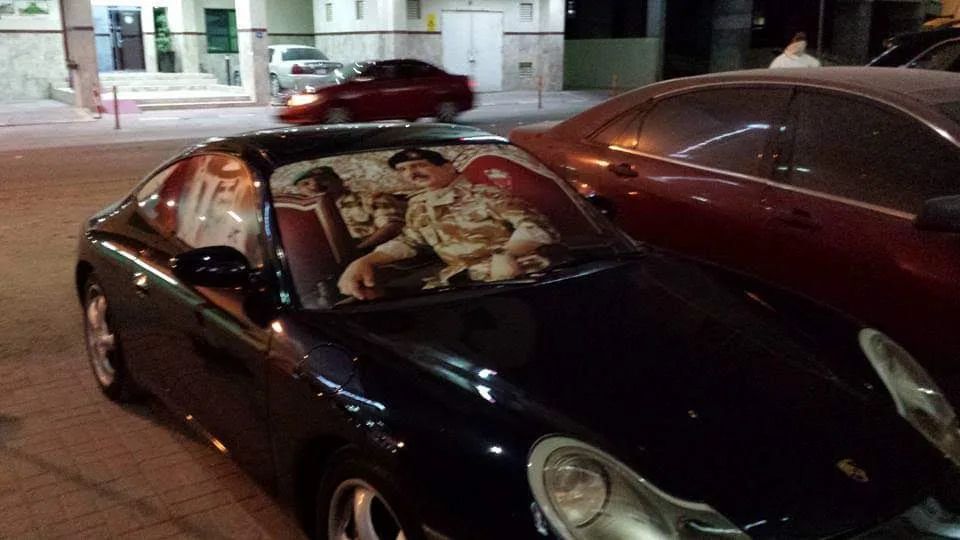 a black sports car parked at night has a large printed image of iraqi president saddam hussein on its windshield, showing…