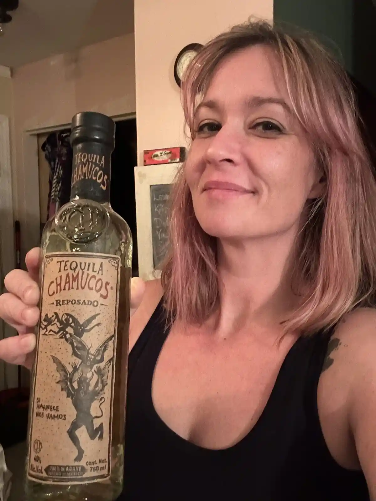 A woman with pinkish hair holds up a bottle of Tequila Chamucos Reposado, which features a demonic figure on its label. She…