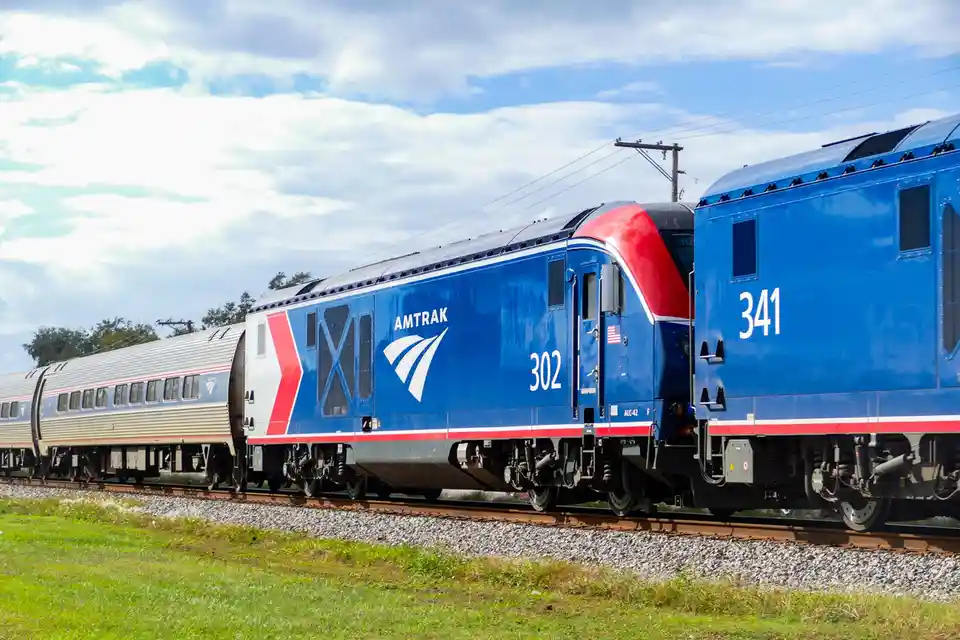 Two Amtrak locomotives, numbered 302 and 341, are shown side-by-side on parallel tracks under a partly cloudy sky, with…