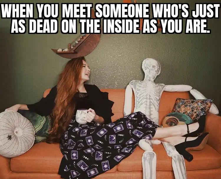 A woman with long red hair sits on an orange couch, smiling while holding a mug, next to a white skeleton also lounging on…