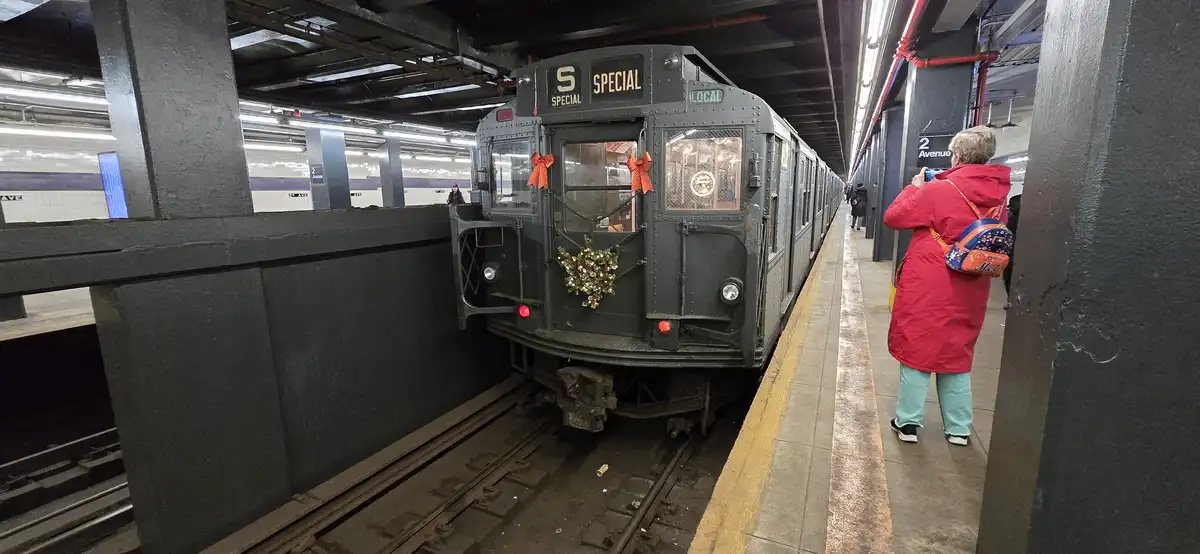 A vintage dark green subway train, marked 'S SPECIAL' and 'LOCAL', is stopped at a station platform decorated with red bows…