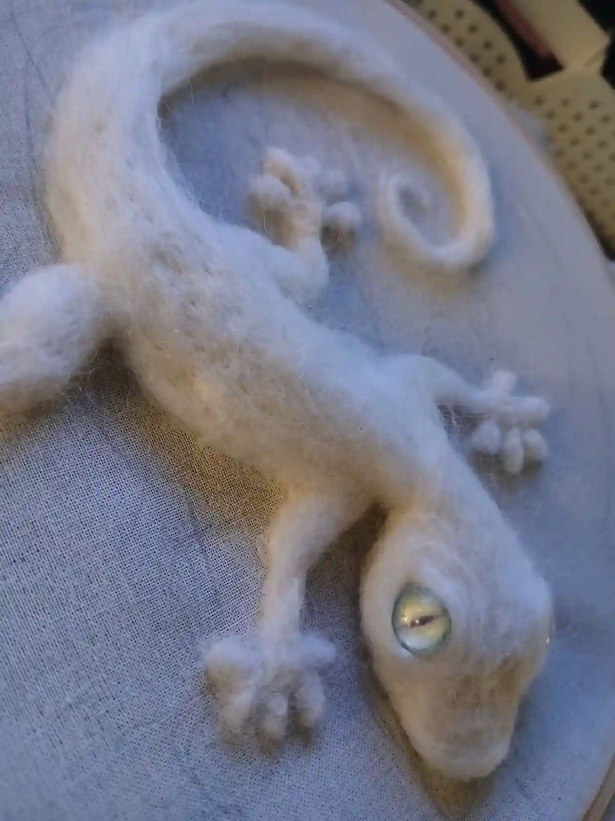 A white, needle-felted gecko sculpture with one large, detailed eye is curled on fabric inside an embroidery hoop. The…