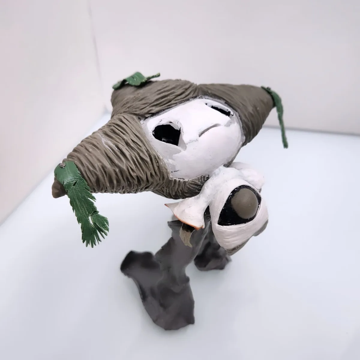 A handmade figurine of a character resembling a stylized, white-faced creature with a brown, textured hat adorned with green…