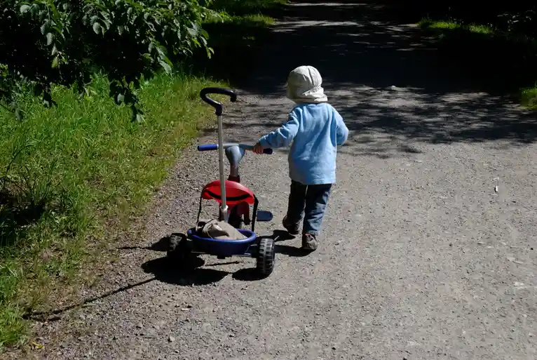 A small child wearing a light blue shirt and a white sun hat walks away from the camera down a gravel path, pushing a red…
