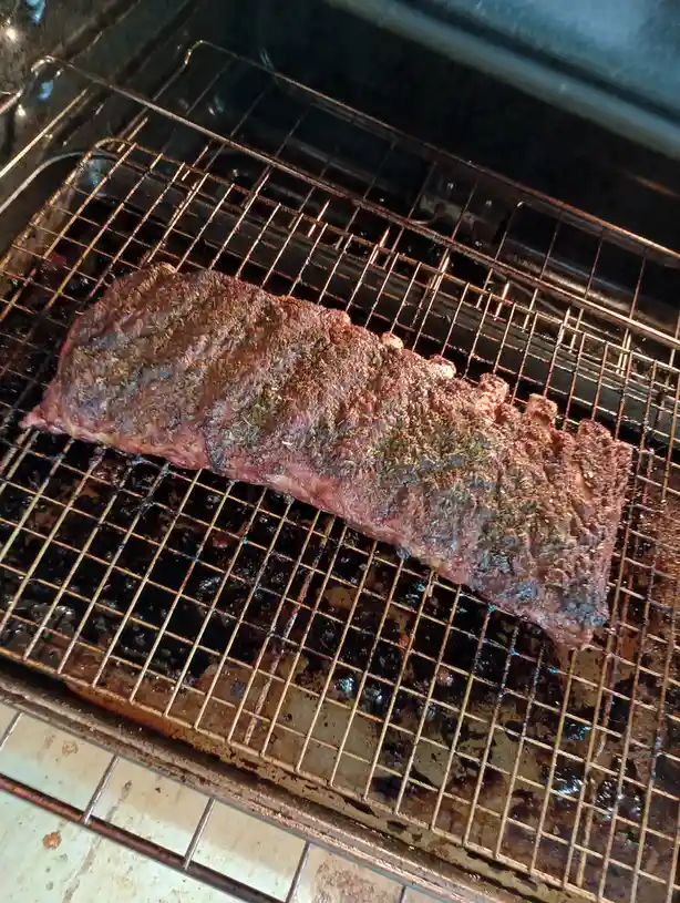 A large rack of seasoned ribs sits on a metal oven rack inside an oven, with visible grill marks and spices on the meat. The…