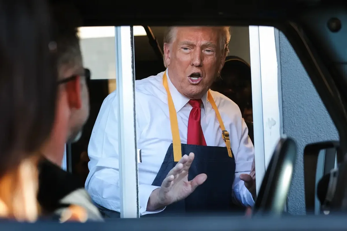 a man wearing a white shirt, red tie, and dark blue apron with yellow straps is leaning out of a vehicle window, gesturing…