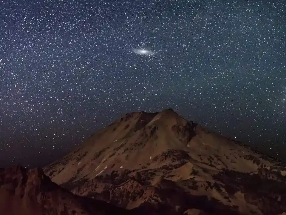 A stunning night photograph shows a mountain peak under a star-filled sky, with the Andromeda galaxy clearly visible as a…