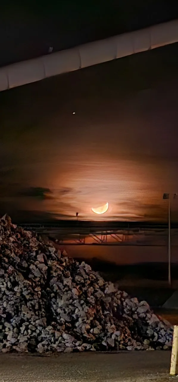 A nighttime photo shows a crescent moon glowing orange above a pile of rubble and industrial structures, with the overlaid…