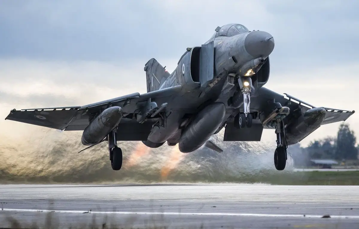 A military jet, likely a Harrier, is captured mid-takeoff with landing gear down, kicking up water spray from a wet runway…