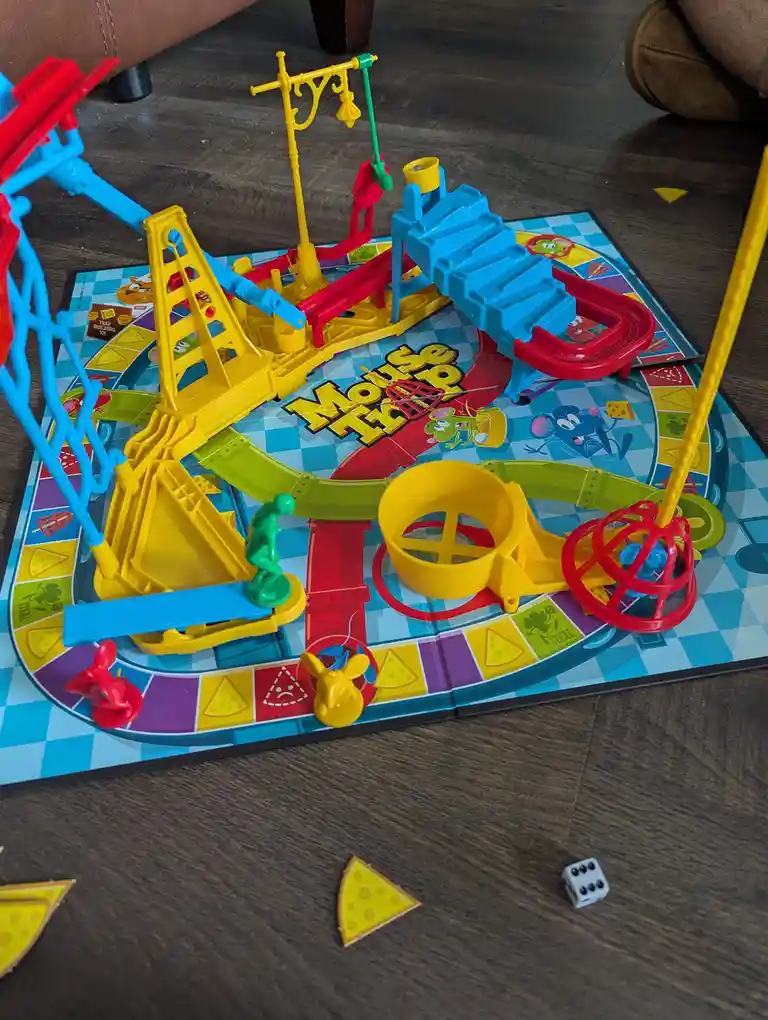 A colorful, partially assembled 'Mouse Trap' board game sits on a wooden floor, featuring bright plastic traps, ramps, and a…