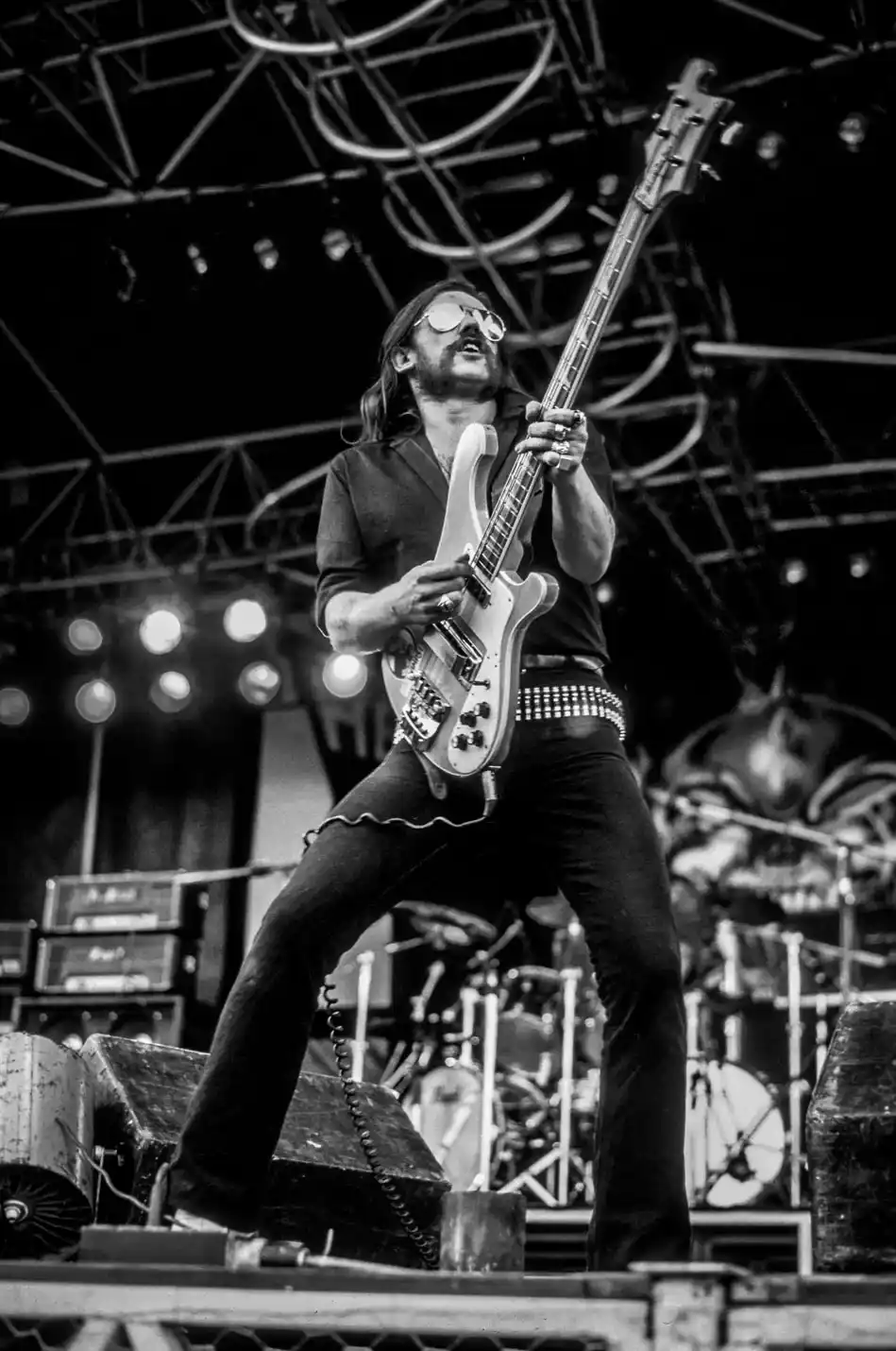 A black and white photo of a man with long hair and sunglasses playing a bass guitar on stage with a dramatic pose. He wears…