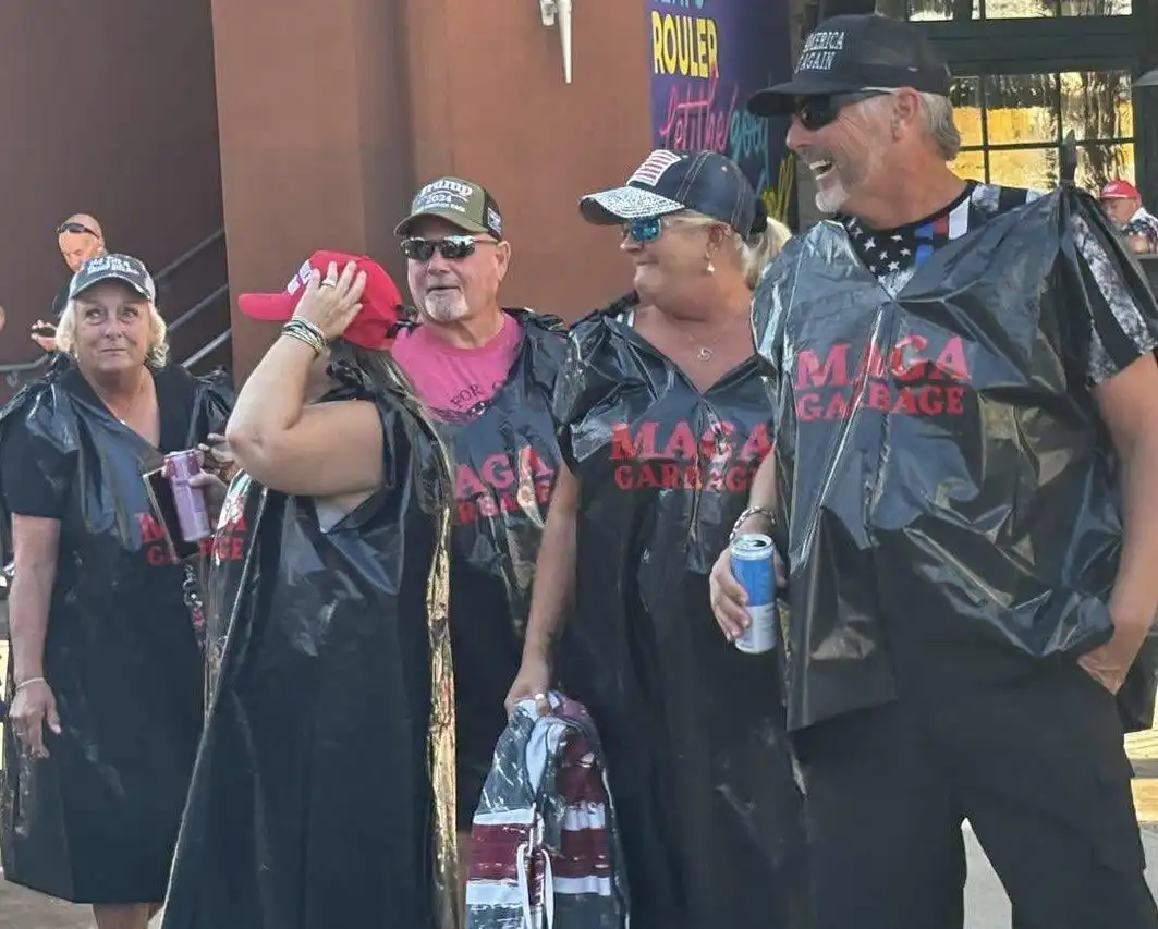 A group of people wearing black trash bag costumes with 'MAGA GARBAGE' printed in red letters on the front, standing…
