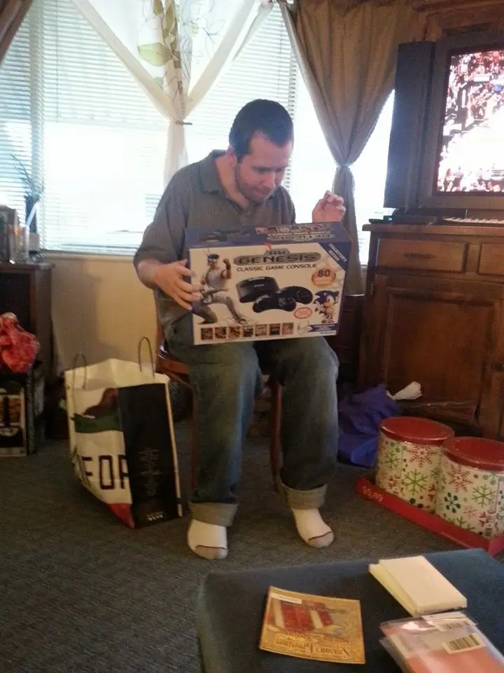 A man sits on a chair in a living room, holding a Sega Genesis Classic Game Console box, looking down at it with a slightly…